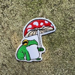 May include: Embroidered patch depicting a green frog sheltering under a red mushroom with white spots. The frog has a yellow and black detail on its head. The patch is outlined in white, suitable for clothing or accessories.