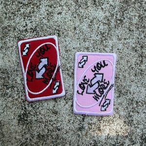 May include: Two embroidered patches resembling Uno reverse cards. One is red with white text reading "Love You More" and the other is pink with the same text. Both have a white border and a reverse arrow symbol.
