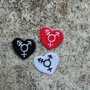 Könnte beinhalten: Drei herzförmige Aufnäher: einer schwarz, einer rot und einer weiß. Jeder Aufnäher zeigt ein Transgender-Symbol in einer Kontrastfarbe. Die Aufnäher sind gestickt und haben einen genähten Rand. Der Hintergrund ist eine strukturierte, graue Oberfläche.