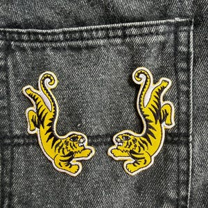 May include: Two embroidered patches featuring yellow tigers with black stripes, depicted in a leaping pose. The tigers have white accents on their bellies and tails, with detailed facial features, positioned against a dark gray denim fabric background.