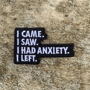 Pode incluir: Patch bordado preto com texto branco: "I CAME. I SAW. I HAD ANXIETY. I LEFT." O patch tem uma forma única, com uma base retangular e uma seção estendida no lado direito.