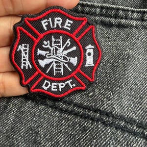 May include: A black and red embroidered patch in the shape of a Maltese cross. The patch features the white text FIRE DEPT and a central emblem depicting a ladder, a fire hydrant, and crossed tools, all outlined in a bold red border.