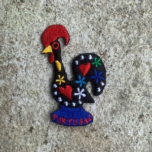 May include: Embroidered patch of a black rooster with a red comb and yellow beak. The rooster is decorated with hearts, flowers, and the text "PORTUGAL" on a blue base. The patch is designed for decorative purposes.