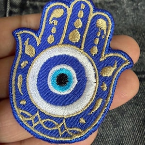 May include: Embroidered Hamsa hand patch with a blue eye design. The patch is blue with gold and white embroidered details. The eye has a black pupil, surrounded by blue and white rings. A decorative item.