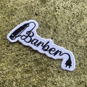 May include: White embroidered patch with the word "Barber" in cursive script, accompanied by a black and white illustration of a hair clipper and a power plug. The patch has a white border.