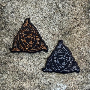 May include: Two triangular embroidered patches. One is black with gold thread, the other is black with silver thread. Each patch features an intricate design of lines and shapes, possibly a symbol or emblem.