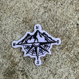 Peut inclure: Un écusson brodé blanc et noir représentant une rose des vents avec des montagnes et les points cardinaux N, E, S et O. L'écusson a une bordure blanche et est destiné aux vêtements ou accessoires.