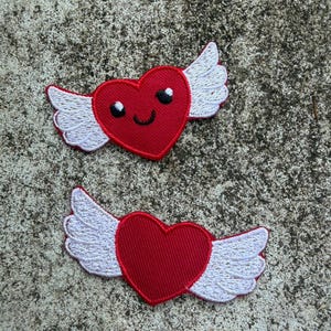 May include: Two embroidered patches featuring red hearts with white wings. One heart has a smiling face with black eyes and a curved mouth. The other heart has no face. Both patches are on a textured gray surface.