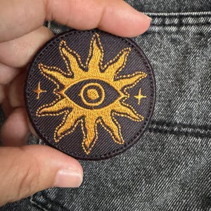 Puede incluir: Un parche bordado circular con un fondo gris oscuro y un diseño de sol en color amarillo dorado. El centro del sol contiene un motivo de ojo, y dos pequeñas estrellas de cuatro puntas están situadas a los lados izquierdo y derecho.