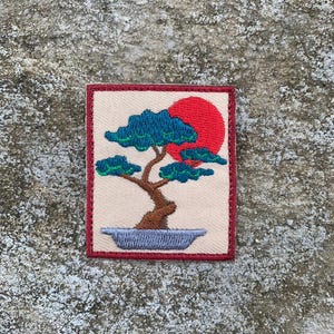 Peut inclure: Écusson brodé représentant un bonsaï avec un feuillage turquoise, un tronc brun et un soleil rouge sur un fond beige. L'écusson est encadré de rouge et posé sur une surface en pierre grise. Le design évoque l'art et la nature japonais.