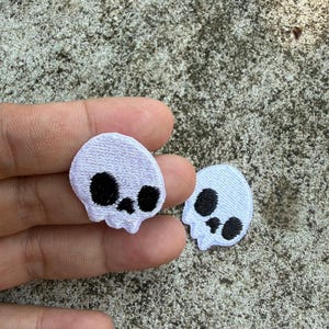 May include: Two small, white skull patches with black eye and nose details. The patches are embroidered and have a textured appearance. One patch is held in a hand, while the other rests on a textured, grey surface.