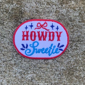 Pode incluir: Um patch oval com fundo branco e borda vermelha. O patch apresenta as palavras "HOWDY Sweetie" em escrita vermelha e azul, com elementos decorativos como um laço e estrelas.