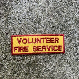 Può includere: Una toppa rettangolare gialla con testo ricamato in rosso che recita "VOLUNTEER FIRE SERVICE". La toppa ha un bordo rosso ed è posta su uno sfondo grigio strutturato.