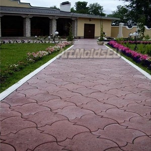 May include: A pathway made of interlocking concrete paving slabs in a reddish-brown colour. The slabs are arranged in a pattern of overlapping arches. The pathway leads to a house with white trim and a green lawn.