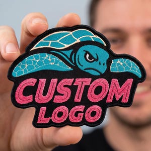 May include: An embroidered patch featuring a teal sea turtle with a black outline and a black background. Below the turtle, the words CUSTOM LOGO are stitched in bright pink thread. The patch has a black border and a distinct, irregular shape.