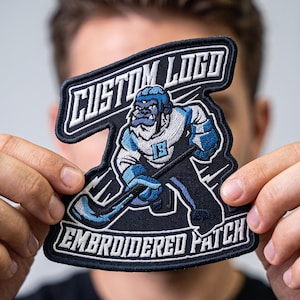 May include: A custom embroidered patch featuring a blue and white gorilla hockey player holding a hockey stick. The patch has a black background with white text that reads CUSTOM LOGO at the top and EMBROIDERED PATCH at the bottom.