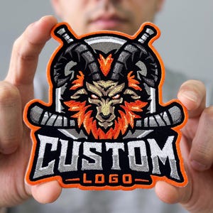 May include: An embroidered patch featuring a central goat head with orange fur and dark horns, framed by two crossed hockey sticks. The design includes a circular grey background and the text CUSTOM LOGO in bold white letters with grey shading.