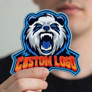 May include: An embroidered patch featuring a stylized white bear head with a blue border and an open mouth showing teeth. Below the bear, the text CUSTOM LOGO is stitched in orange with a red outline, set against a dark blue background.