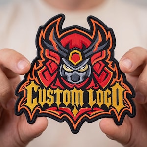 May include: An embroidered patch featuring a stylized warrior mask in red, orange, and black with sharp, angular lines. The text "CUSTOM LOGO" is stitched in bold, yellow, gothic-style lettering across the bottom of the design.