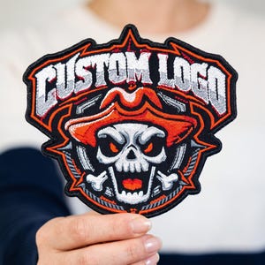 May include: An embroidered patch featuring a skull wearing an orange hat, with the text CUSTOM LOGO arched above in white and grey. The design includes black, orange, and white thread, with a black border and sharp, angular edges.
