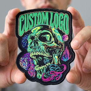 May include: A black embroidered patch featuring the text CUSTOM LOGO in bright green thread at the top. Below the text is a detailed skull illustration rendered in shades of neon green, blue, and yellow, surrounded by purple and pink abstract shapes.