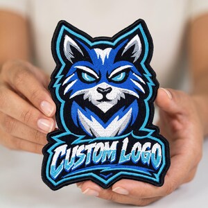 May include: An embroidered patch featuring a stylized blue and white wolf head mascot with glowing light blue eyes. Below the wolf head, the text CUSTOM LOGO is stitched in a bold, white, and light blue font against a black background.
