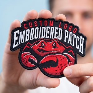 May include: A black embroidered patch featuring a red crab graphic with white text that reads CUSTOM LOGO EMBROIDERED PATCH. The patch has a scalloped edge and is stitched with red and white thread on a black fabric background.