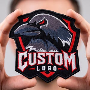 May include: A shield-shaped embroidered patch featuring a black raven head with red accents above a smaller wolf head. The text CUSTOM LOGO is stitched in white with red outlines at the bottom. The patch has a black border and red geometric details.