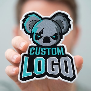 May include: An embroidered patch featuring a stylized gray koala head with teal accents above the text CUSTOM LOGO in teal and gray block lettering. The design is outlined in white and set against a black background with a thick white border.