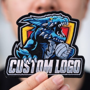 May include: An embroidered patch featuring a blue dragon mascot holding a volleyball in front of a yellow and red shield emblem. The text CUSTOM LOGO is stitched in white block letters on a black background at the bottom of the patch design.