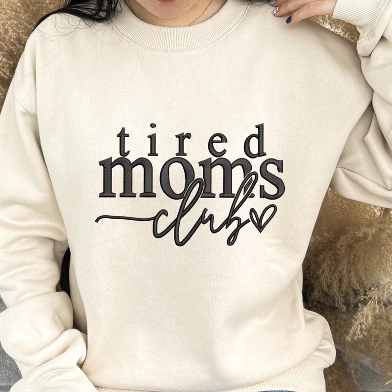 Peut inclure: Un sweat-shirt cr&egrave;me avec le texte "tired moms club" en lettres cursives noires avec un petit c&oelig;ur.