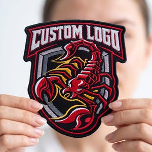 May include: A shield-shaped embroidered patch featuring a red and yellow scorpion graphic centered on a black background. The top of the patch displays the text CUSTOM LOGO in white block letters with a red outline, framed by a black border.