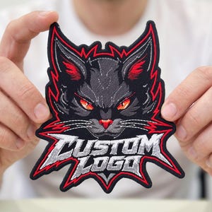 May include: An embroidered patch featuring a stylized black cat head with sharp, pointed ears, glowing orange eyes, and a red nose. The design is outlined in vibrant red thread and includes the text CUSTOM LOGO in a jagged, silver-grey font.