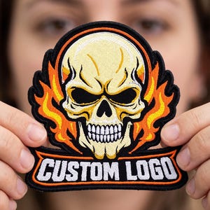 May include: An embroidered patch featuring a central cream-colored skull surrounded by orange and yellow flames. Below the skull, a black banner with an orange border contains the white text CUSTOM LOGO in a bold, sans-serif capital font.