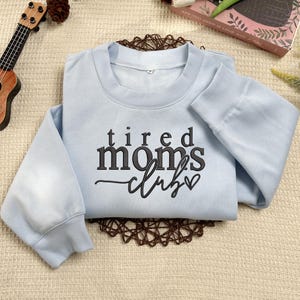 Peut inclure: Sweat-shirt bleu clair avec le texte "tired moms club" en lettres noires avec un petit c&oelig;ur.