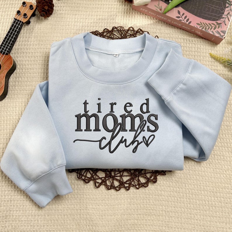 Peut inclure: Sweat-shirt bleu clair avec le texte "tired moms club" en lettres noires avec un petit c&oelig;ur.