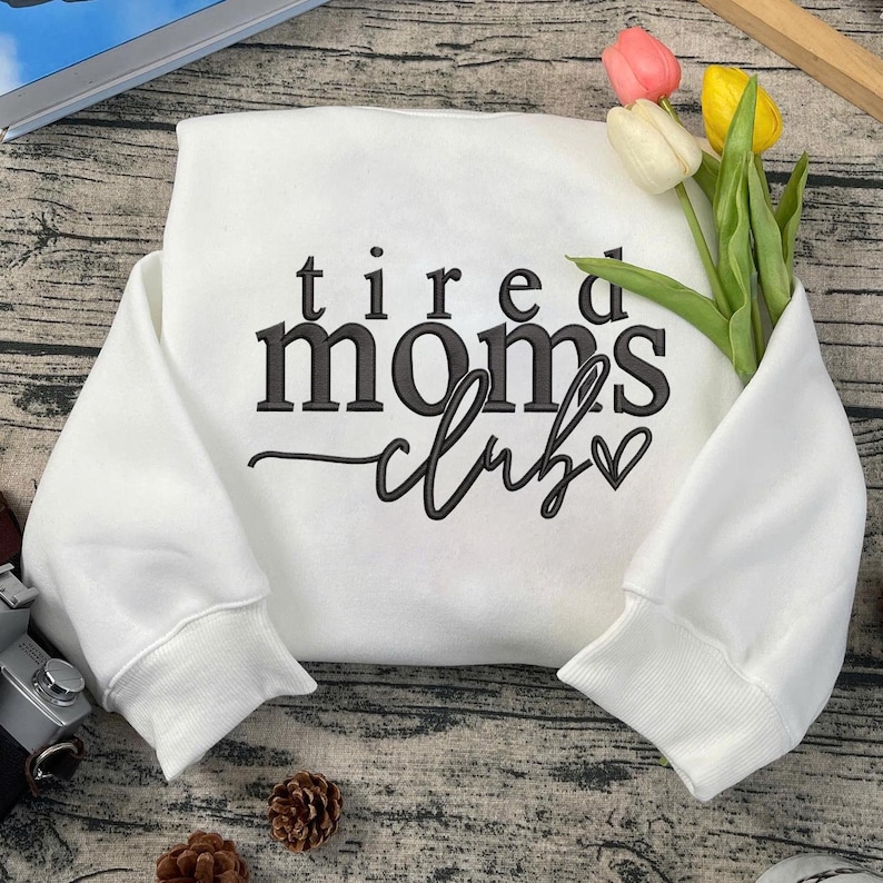 Peut inclure: Sweat-shirt blanc avec un texte noir qui dit "tired moms club" avec un petit c&oelig;ur.