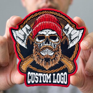 May include: A circular embroidered patch featuring a skull with a red beanie, brown beard, and glasses, flanked by two crossed axes. The design is set against a rope-like border with a red edge and the text CUSTOM LOGO in white at the bottom.