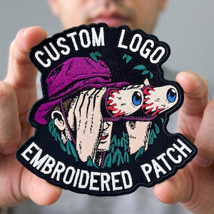 May include: A black embroidered patch featuring a person wearing a purple bucket hat and looking through binoculars that are shaped like two large eyeballs with blue irises. The text CUSTOM LOGO is at the top and EMBROIDERED PATCH is at the bottom.