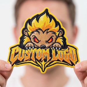 May include: An embroidered patch featuring a stylized, spiky-haired creature with red eyes and sharp claws. The design includes the text CUSTOM LOGO in bold, jagged yellow and orange lettering, all outlined in a thick yellow border on a black background.