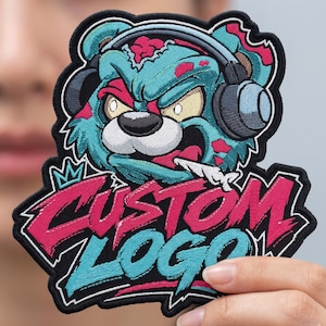 May include: An embroidered patch featuring a teal bear character wearing grey headphones with a fierce expression. Below the bear, the text CUSTOM LOGO is stitched in bold pink and teal letters with a black border and a small crown icon above.