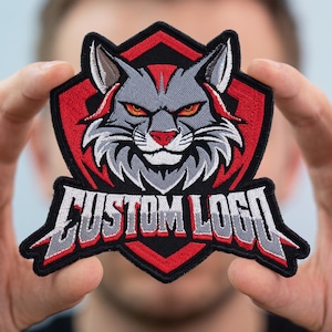 May include: An embroidered patch featuring a stylized gray bobcat head with orange eyes and red accents, set against a black and red shield-shaped background. Below the animal head, the text CUSTOM LOGO is stitched in bold gray and red letters.