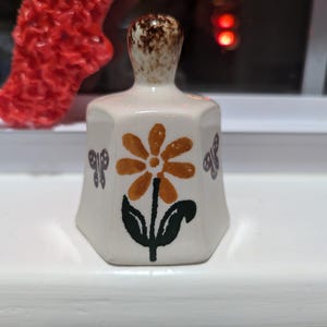 May include: A small white ceramic bell with a brown top. The bell is decorated with a yellow flower with orange petals and green leaves. Two gray butterflies are on either side of the flower.