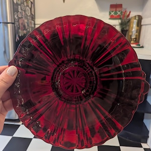 May include: A vibrant red glass plate with a fluted design and scalloped edges. The plate has a starburst pattern in the center and is held by a person. The plate is likely vintage or antique.