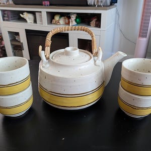 May include: A ceramic tea set consisting of a teapot with a woven rattan handle and four matching cups. The pieces feature an off-white speckled glaze with a prominent horizontal yellow band around the base and a thin dark brown accent line.