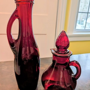 May include: Two pieces of ruby red glass serveware. A tall, slender pitcher with a curved handle and a small, round decanter with a handle and a stopper. The pitcher is approximately 12 inches tall, and the decanter is about 6 inches tall.