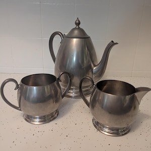 May include: A vintage pewter tea set, including a teapot, a sugar bowl, and a creamer. The set has a classic design with curved handles and a shiny, metallic finish. The teapot has a domed lid and a long spout. The sugar bowl and creamer have rounded bodies.