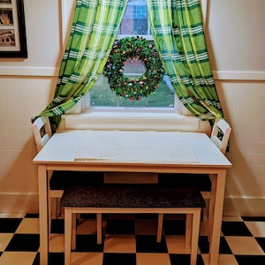 May include: A white table with two chairs and a bench, set against a black and white checkered floor. A green and white plaid curtain hangs in the window behind the table. A Christmas wreath with lights hangs in the window.