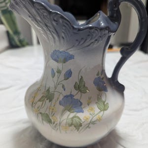 May include: A blue and white ceramic pitcher with a floral design. The pitcher has a wide mouth and a handle. The flowers are blue, yellow, and green.