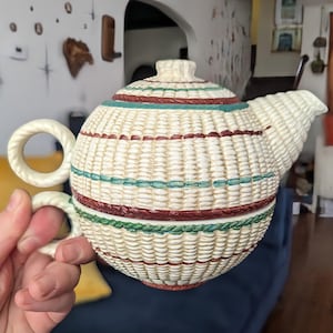 May include: A ceramic teapot with a textured, cream-colored basket-weave pattern. The body features horizontal bands in shades of teal, dark red, and green. It has a round handle, a short spout, and a matching lid with a small knob on top.